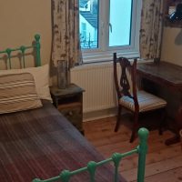 Photo of iron bedstead, wooden floorboards, with small desk and window